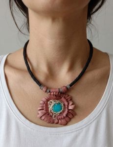 A handcrafted bohemian choker necklace displayed on a woman's neck, featuring a flower design made of pink-brown leather petals with a blue turquoise stone in the center and a black braided cord.